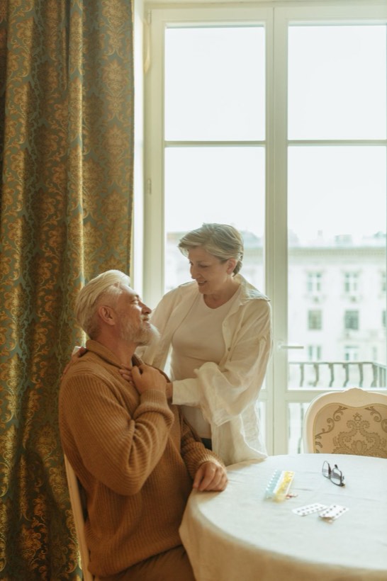 Seniors pie galda ar medikamentiem, blakus stāv tuviniece
